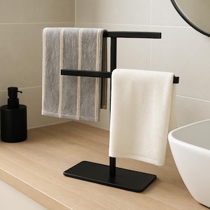 Pode incluir: Um toalheiro de metal preto com duas toalhas. Uma é branca, a outra cinza com listras verticais. Um dispensador de sabonete preto está na bancada, em um banheiro.