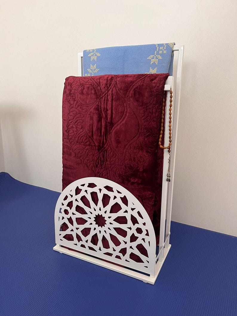May include: White metal prayer rug stand with a decorative cut-out design. Two prayer rugs are folded and stored on the stand. A set of prayer beads hangs from the stand.