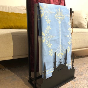 May include: A black metal prayer rug stand with a mosque silhouette design. It has two prayer rugs hanging on it, one red and one light blue with white embroidery.