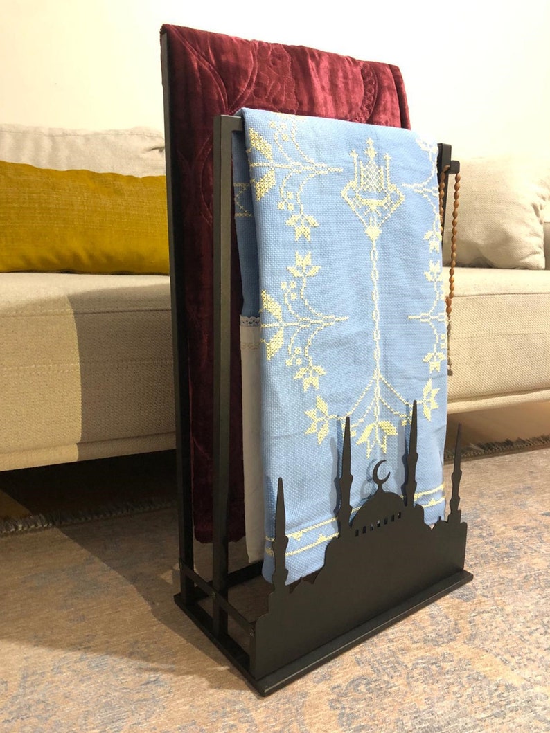 May include: A black metal prayer rug stand with a mosque silhouette design. It has two prayer rugs hanging on it, one red and one light blue with white embroidery.