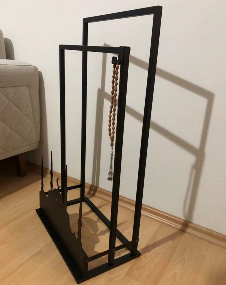 May include: Black metal stand with a silhouette of a mosque and a crescent moon. A wooden prayer bead necklace hangs from the top of the stand.