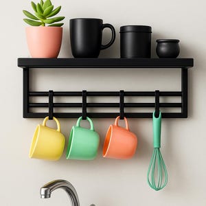 Puede incluir: Estante de cocina de metal negro con tazas colgantes amarillas, verdes y naranjas. Un batidor verde cuelga junto a las tazas. Una maceta rosa con una planta verde y tazas y recipientes negros están en el estante.