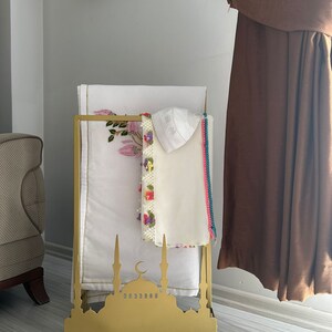 May include: A gold metal stand shaped like a mosque with a crescent moon on top. The stand has two shelves for holding prayer rugs and a white prayer cap.