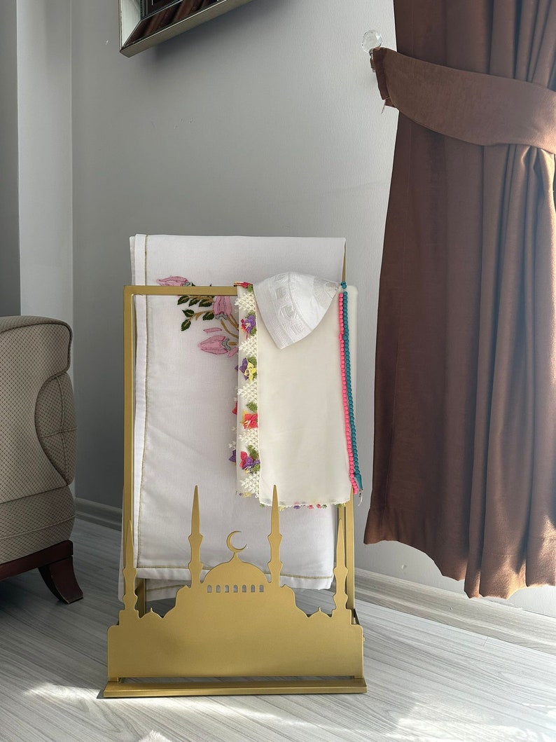 May include: A gold metal stand shaped like a mosque with a crescent moon on top. The stand has two shelves for holding prayer rugs and a white prayer cap.
