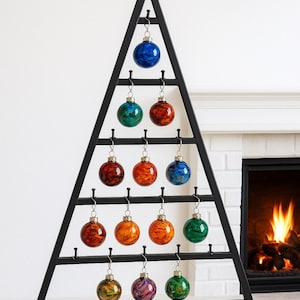 May include: A black metal Christmas tree-shaped ornament display holding 15 colorful glass ball ornaments. The ornaments are in shades of blue, green, orange, red, and purple. A fireplace with a burning fire is in the background.