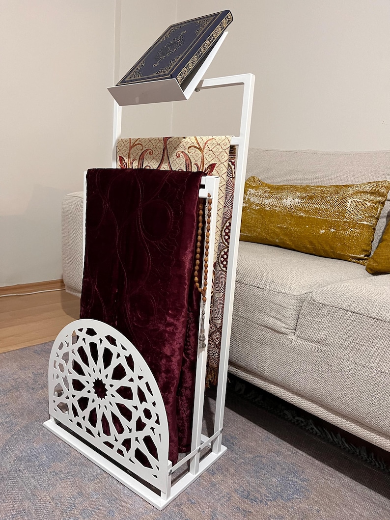 May include: White metal prayer stand with a burgundy prayer rug, a patterned prayer rug, and a book on top. The stand has a decorative cut-out design at the bottom.