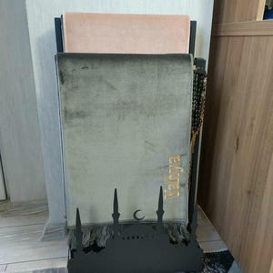 May include: A black metal prayer rug stand with a mosque design. Two prayer rugs are stacked on the stand, one is pink and the other is gray. The gray rug has the word "Yahya" embroidered on it in gold thread.
