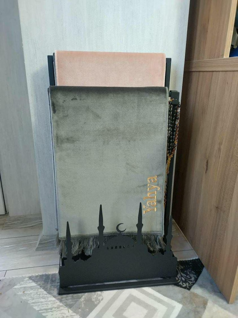 May include: A black metal prayer rug stand with a mosque design. Two prayer rugs are stacked on the stand, one is pink and the other is gray. The gray rug has the word "Yahya" embroidered on it in gold thread.
