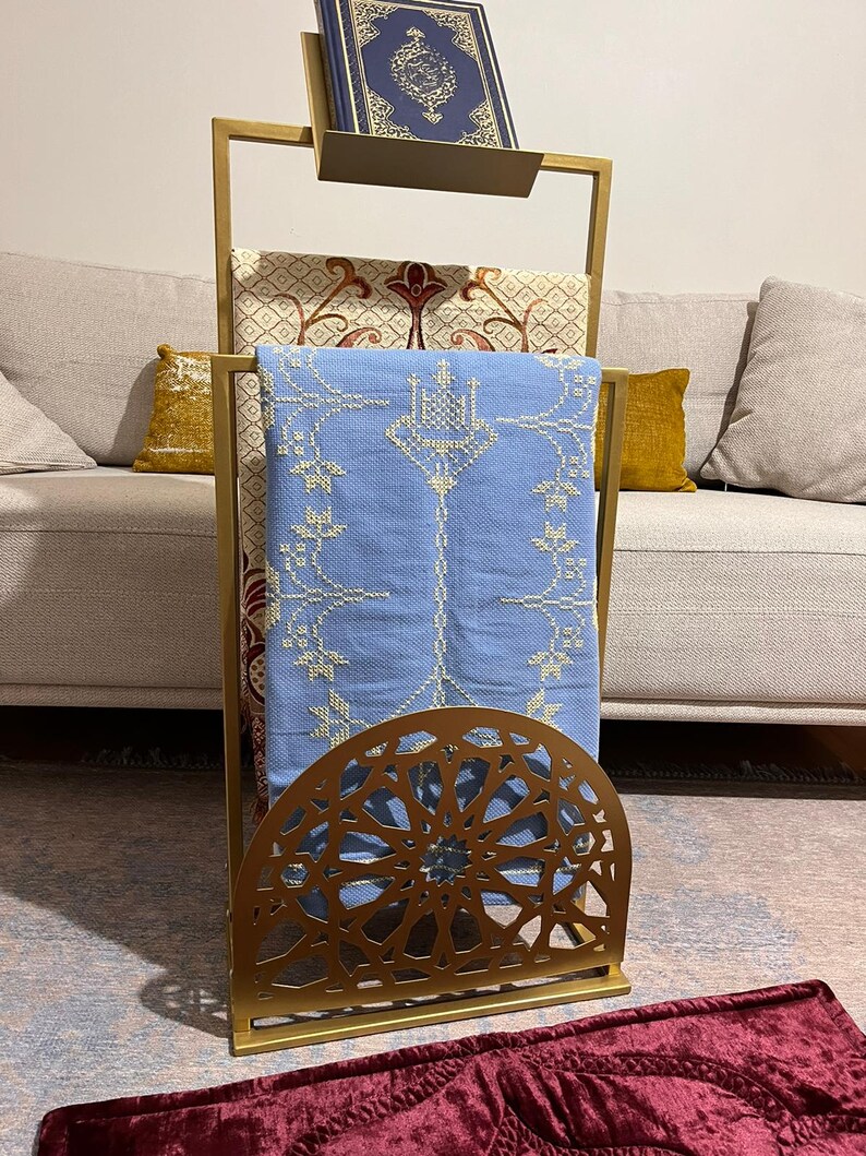 May include: A gold metal prayer stand with a decorative design. The stand has two shelves for holding prayer rugs and a book. The stand is on a red rug.