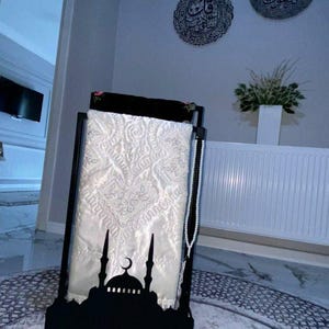 May include: A black metal prayer rug stand with a white prayer rug and a string of white beads. The stand features a silhouette of a mosque with a crescent moon.