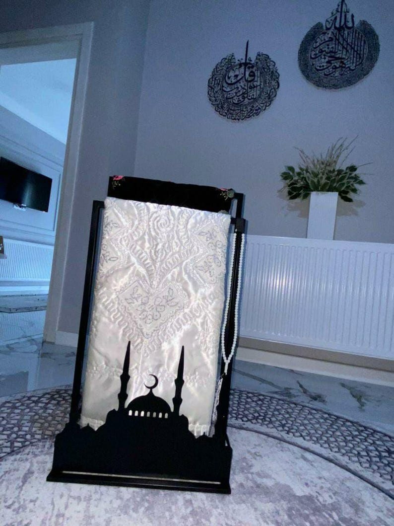 May include: A black metal prayer rug stand with a white prayer rug and a string of white beads. The stand features a silhouette of a mosque with a crescent moon.