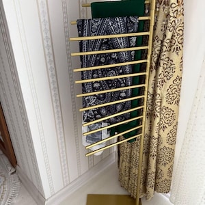 May include: A gold metal stand with multiple horizontal bars for holding scarves. The stand has a rectangular base and is decorated with a paisley pattern.