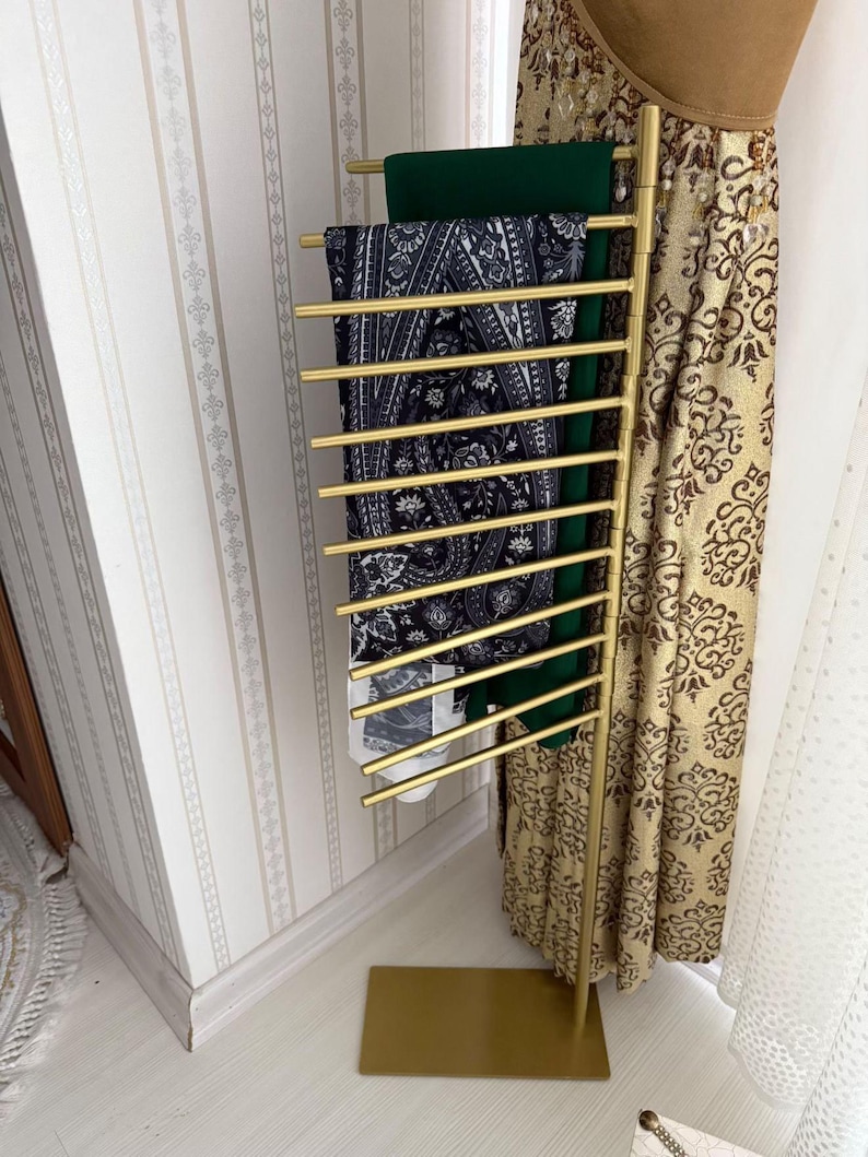 May include: A gold metal stand with multiple horizontal bars for holding scarves. The stand has a rectangular base and is decorated with a paisley pattern.