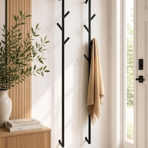 Puede incluir: Dos percheros de metal negro montados en una pared blanca. Cada perchero tiene múltiples ganchos en ángulo. Una manta beige cuelga de un perchero. Los percheros están cerca de un mueble de madera, un jarrón con vegetación y una puerta.