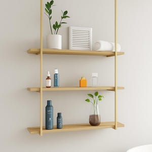 May include: A modern, three-tiered wooden shelf unit suspended from a gold-colored metal frame. The shelves hold various bathroom accessories, including plants, bottles, and a rolled towel. The unit is against a white wall.