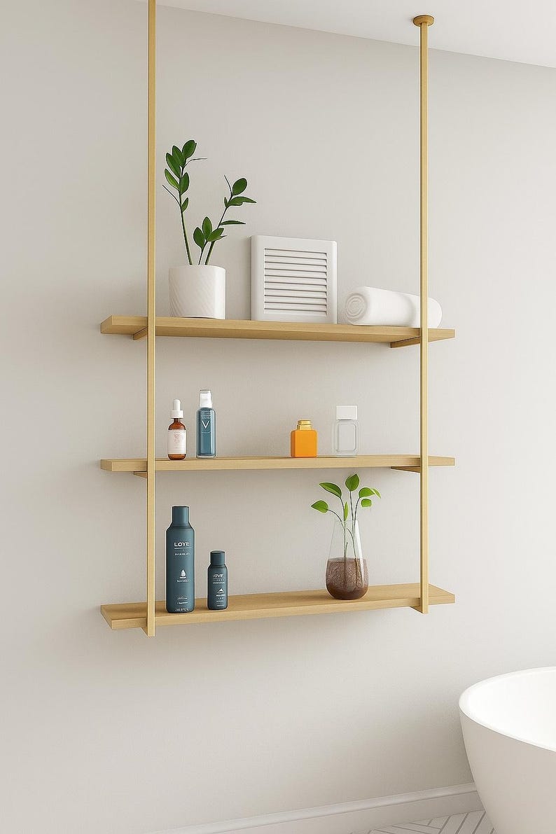 May include: A modern, three-tiered wooden shelf unit suspended from a gold-colored metal frame. The shelves hold various bathroom accessories, including plants, bottles, and a rolled towel. The unit is against a white wall.