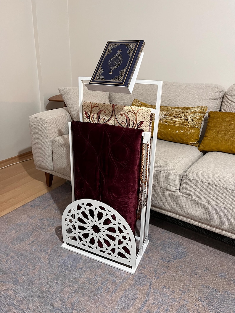 May include: White metal prayer stand with a decorative cut-out design. The stand holds a prayer rug, a second rug, and a book.