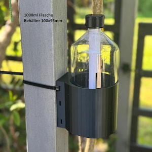Op de afbeelding: Een doorzichtige plastic fles met een zwarte dop wordt vastgehouden in een zwarte houder die aan een grijze paal is bevestigd. De fles is gelabeld met "1000ml Flasche Behälter 100x95mm". De houder is gemonteerd met een zwarte tie-wrap.