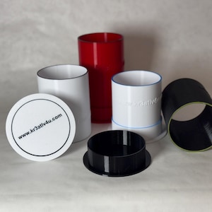 Op de afbeelding: Een assortiment cilindrische containers en een deksel. De containers zijn wit, rood en zwart, sommige met gekleurde randen. Eén deksel is wit met de tekst "www.kr3ativ4u.com" in zwart.