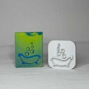 May include: A rectangular bar of soap with a gradient green and blue finish featuring a blue outline of a clawfoot bathtub with bubbles, alongside a white plastic cookie cutter shaped like a clawfoot bathtub with rising bubbles.