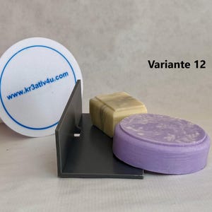 Pode incluir: Um saboneteira de plástico cinza com duas barras de sabão. Uma barra é de cor verde claro e a outra é de cor roxo claro. A saboneteira tem o texto "Variante 12" impresso nela.