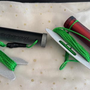 Puede incluir: Dos carretes de hilo verde con soportes de plástico blanco. Un carrete está unido a un cilindro negro con el texto "Pellingbergetool" impreso en él. El otro carrete está unido a un cilindro rojo con el texto "All-Batik" impreso en él.