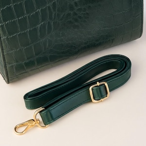 A dark green leather handbag with a gold-colored metal strap and buckle is placed on a white surface.