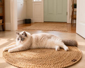 May include: A circular, braided jute pet bed in light brown, with a small tag in the corner. A fluffy, light-coloured cat with blue eyes is lying on the bed. The bed is on a tiled floor. The bed measures approximately 90 cm in diameter.