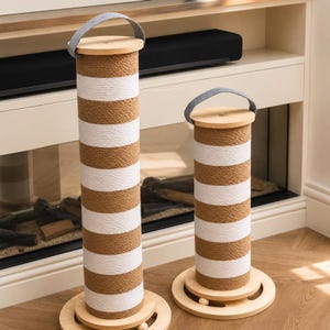 May include: Two cat scratching posts with a striped design. The posts are cylindrical, wrapped in natural-coloured rope with white stripes. Each post has a wooden top and base, and a grey handle. The posts are different sizes.
