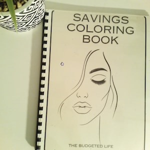 May include: A black and white colouring book with the title "Savings Coloring Book" and the text "The Budgeted Life" at the bottom. The cover features a line drawing of a woman's face with her eyes closed.