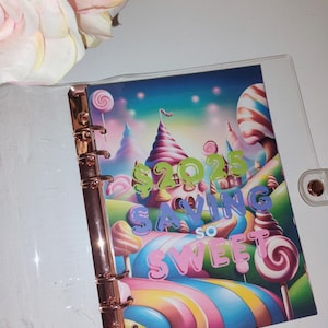 May include: A clear plastic binder with a colorful image of a candy land themed landscape. The image includes a castle, candy canes, lollipops, and the text "2023 Saving So Sweet".