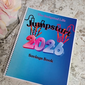 Puede incluir: Un libro de ahorros encuadernado en espiral titulado "Jumpstart 2026" con una cubierta azul degradada. La portada presenta el texto "The Budgeted Life" y "Savings Book". Los números "2026" tienen un degradado rosa y azul. Una flor rosa y blanca está en el fondo.