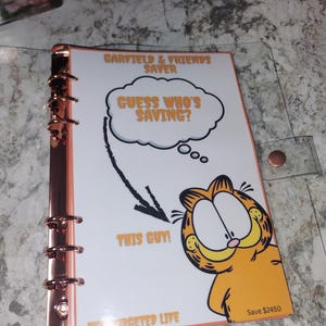 May include: A clear binder with rose gold hardware holds a white page featuring Garfield the cat. The page reads "GARFIELD & FRIENDS SAVER" and "GUESS WHO'S SAVING?" with an arrow pointing to Garfield and the text "THIS GUY!" The bottom reads "THE BUDGETED LIFE".