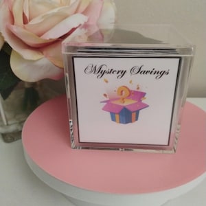 Op de afbeelding: Een transparante acrylbox met een wit label waarop "Mystery Savings" staat. Het label heeft een cartoonillustratie van een kleurrijke doos met een vraagteken erin.