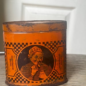 Puede incluir: Una lata de metal naranja con un patrón de cuadros negros y blancos. La lata tiene una imagen de una mujer con un vestido blanco y negro sosteniendo una taza. El texto en la lata dice "GEDEPONEERD MERK" y "GEBRUIKSMODELL".