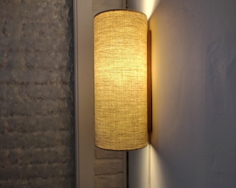 May include: A cylindrical wall sconce featuring a textured, light beige fabric shade mounted on a wooden base. The lamp emits a warm, soft glow from the top and bottom, casting light onto the wall surface behind it.