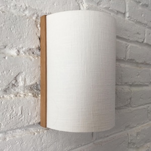Op de afbeelding: Een cilindrische wandlamp van witte stof met een houten rand. De lamp is gemonteerd op een witte bakstenen muur.