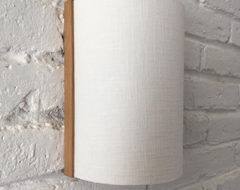 May include: A white fabric cylindrical wall sconce with a wood trim. The sconce is mounted on a white brick wall.
