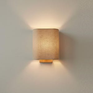 Può includere: Un'applique da parete con una base rettangolare in legno e un paralume cilindrico. Il paralume è in tessuto beige chiaro e l'apparecchio proietta un bagliore caldo sulla parete. L'apparecchio è montato su una parete di colore chiaro.