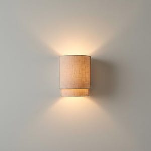 Könnte beinhalten: Beige zylindrische Wandleuchte mit zweistufigem Design. Die Lampe strahlt ein warmes Licht aus, das nach oben und unten scheint. Der Lampenschirm hat eine strukturierte Optik und verleiht der Leuchte Eleganz. Die Leuchte ist an einer neutralfarbenen Wand befestigt.