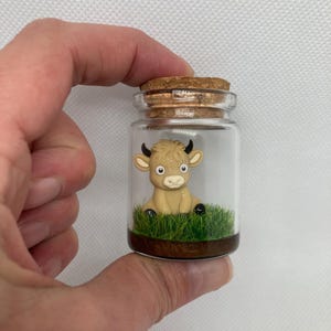 K&ouml;nnte beinhalten: Ein kleines Glasgef&auml;&szlig; mit einem Korkverschluss enth&auml;lt eine Miniaturfigur einer braunen Kuh, die auf gr&uuml;nem Gras sitzt.