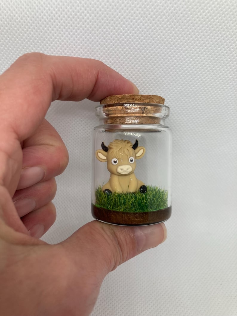 K&ouml;nnte beinhalten: Ein kleines Glasgef&auml;&szlig; mit einem Korkverschluss enth&auml;lt eine Miniaturfigur einer braunen Kuh, die auf gr&uuml;nem Gras sitzt.