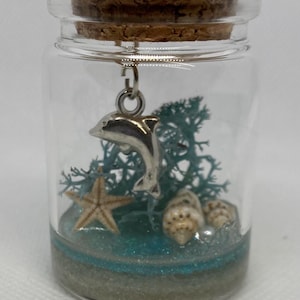 May include: A small glass jar with a cork lid contains a miniature seascape. A silver dolphin charm hangs from the top of the jar. Inside the jar, there is a small starfish, seashells, and blue glitter that resembles sand.