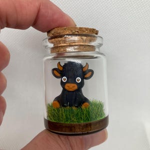 K&ouml;nnte beinhalten: Ein kleines Glasgef&auml;&szlig; mit einem Korkverschluss enth&auml;lt eine schwarze Miniatur-Stierfigur, die auf gr&uuml;nem Moos sitzt.