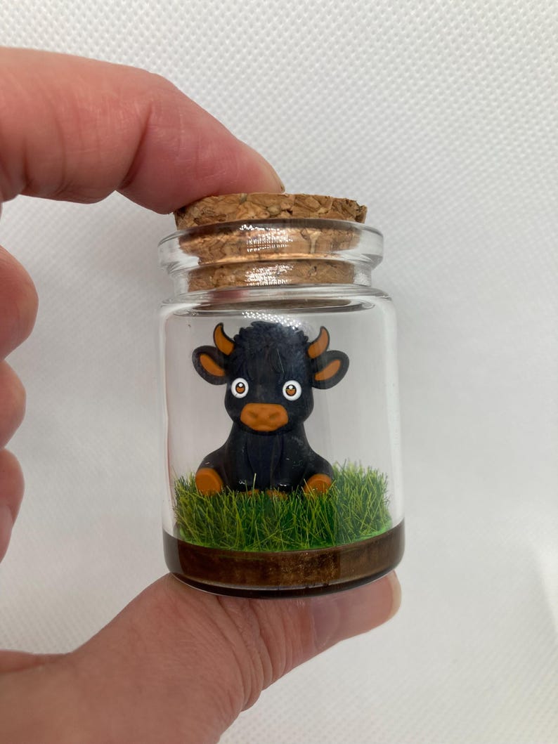 K&ouml;nnte beinhalten: Ein kleines Glasgef&auml;&szlig; mit einem Korkverschluss enth&auml;lt eine schwarze Miniatur-Stierfigur, die auf gr&uuml;nem Moos sitzt.