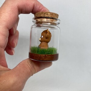 K&ouml;nnte beinhalten: Ein kleines Glasgef&auml;&szlig; mit einem Korkverschluss enth&auml;lt eine Miniaturfigur einer braunen Kuh, die in einem Bett aus gr&uuml;nem Gras steht.