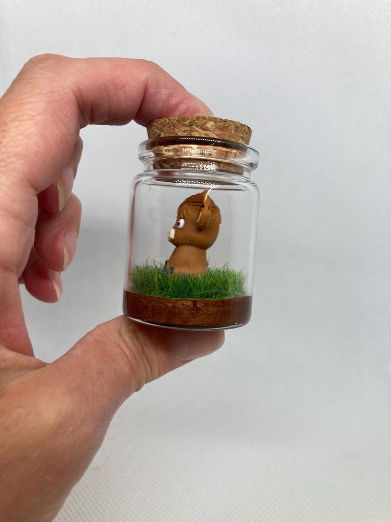 K&ouml;nnte beinhalten: Ein kleines Glasgef&auml;&szlig; mit einem Korkverschluss enth&auml;lt eine Miniaturfigur einer braunen Kuh, die in einem Bett aus gr&uuml;nem Gras steht.
