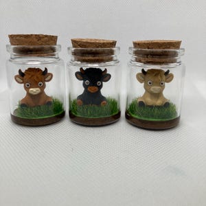 K&ouml;nnte beinhalten: Drei Glasgl&auml;ser mit Korkdeckeln, jedes mit einer kleinen Tonfigur einer Kuh in verschiedenen Farben: braun, schwarz und wei&szlig;. Die K&uuml;he sitzen auf einem Bett aus gr&uuml;nem Gras.