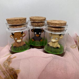 K&ouml;nnte beinhalten: Drei Glasgl&auml;ser mit Korkdeckeln, jedes mit einer kleinen Tonfigur einer Kuh. Die K&uuml;he sind braun, schwarz und wei&szlig;. Die Gl&auml;ser sind mit gr&uuml;nem Gras gef&uuml;llt.