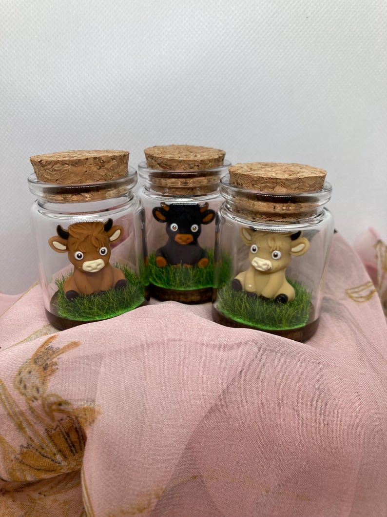 K&ouml;nnte beinhalten: Drei Glasgl&auml;ser mit Korkdeckeln, jedes mit einer kleinen Tonfigur einer Kuh. Die K&uuml;he sind braun, schwarz und wei&szlig;. Die Gl&auml;ser sind mit gr&uuml;nem Gras gef&uuml;llt.